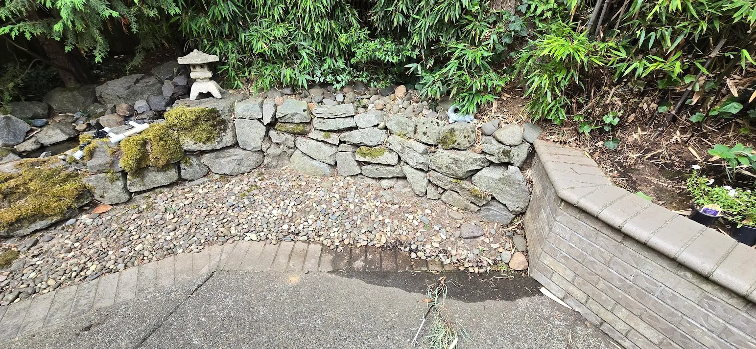 Mossy rock wall with bare gravel and exposed pond plumbing before the rebuild