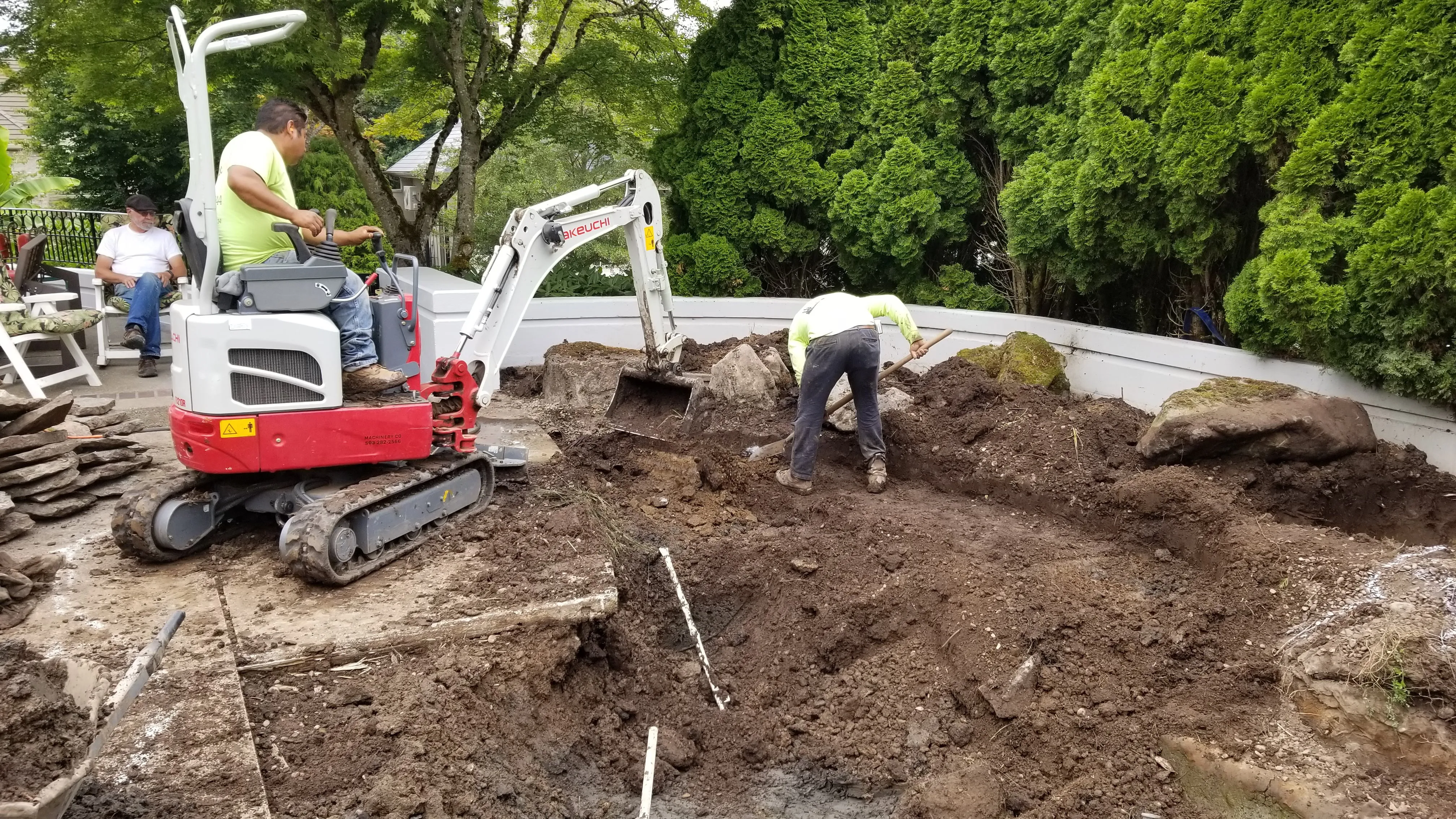 Mini excavator and crew digging the pond basin against the backyard wall