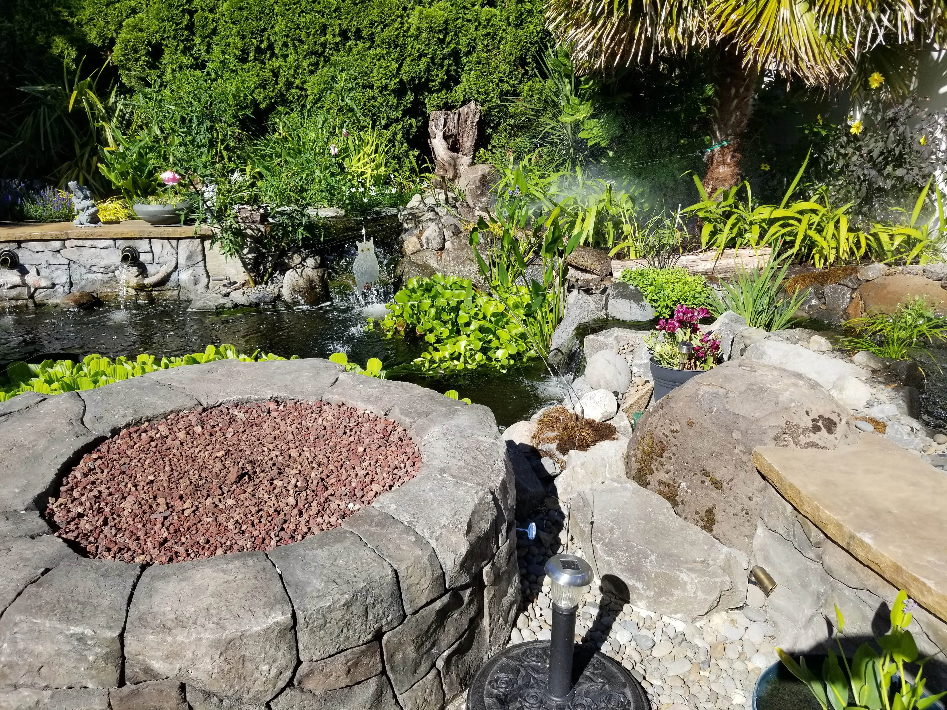Carved concrete fire pit filled with red lava rock at the edge of the koi pond