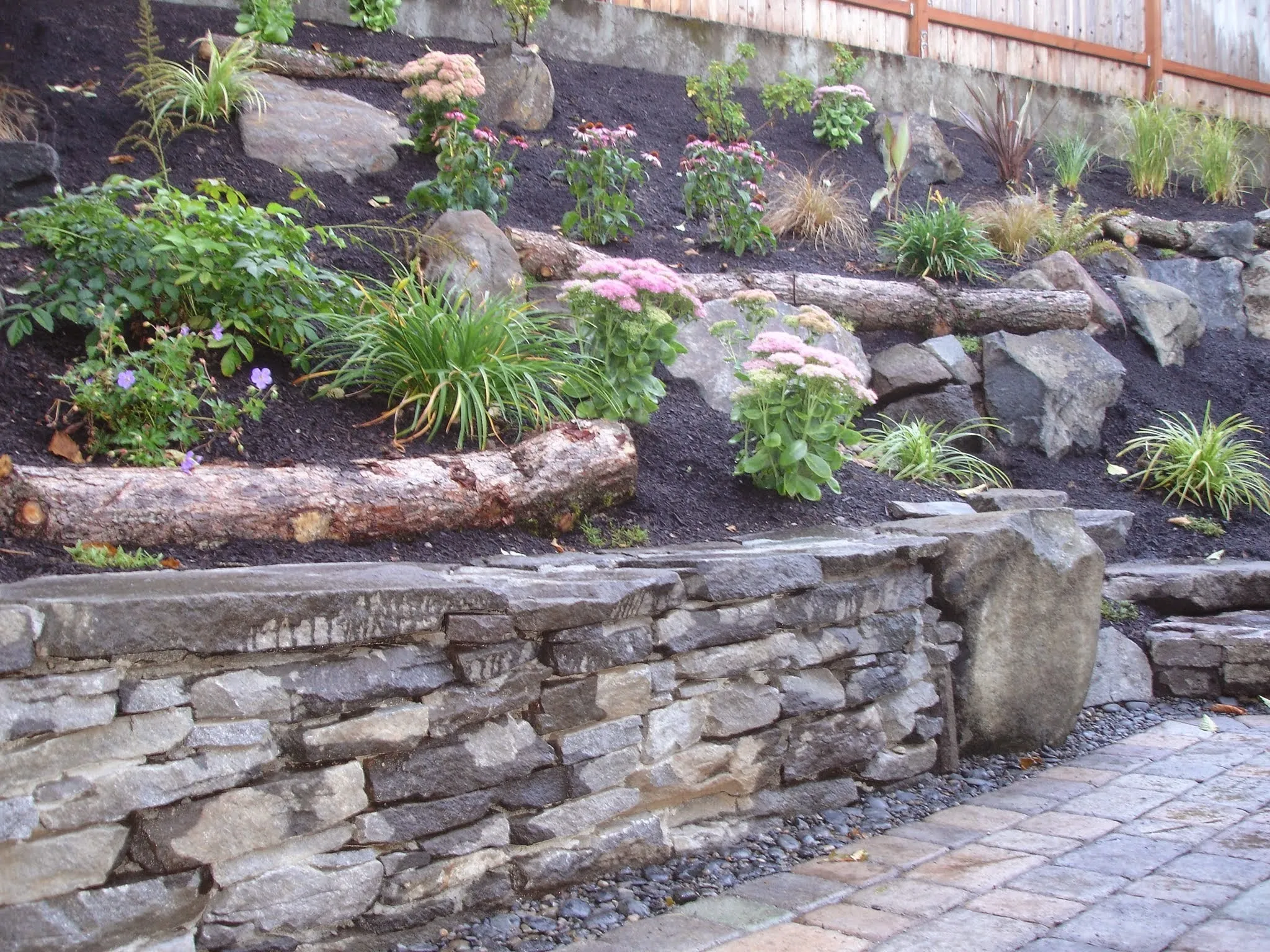 Dry-stacked basalt retaining wall holding a terraced hillside planting bed above a paver path